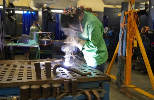 A student wearing a welding helmet and heavy gloves uses a welder on a metal piece in a workshop.