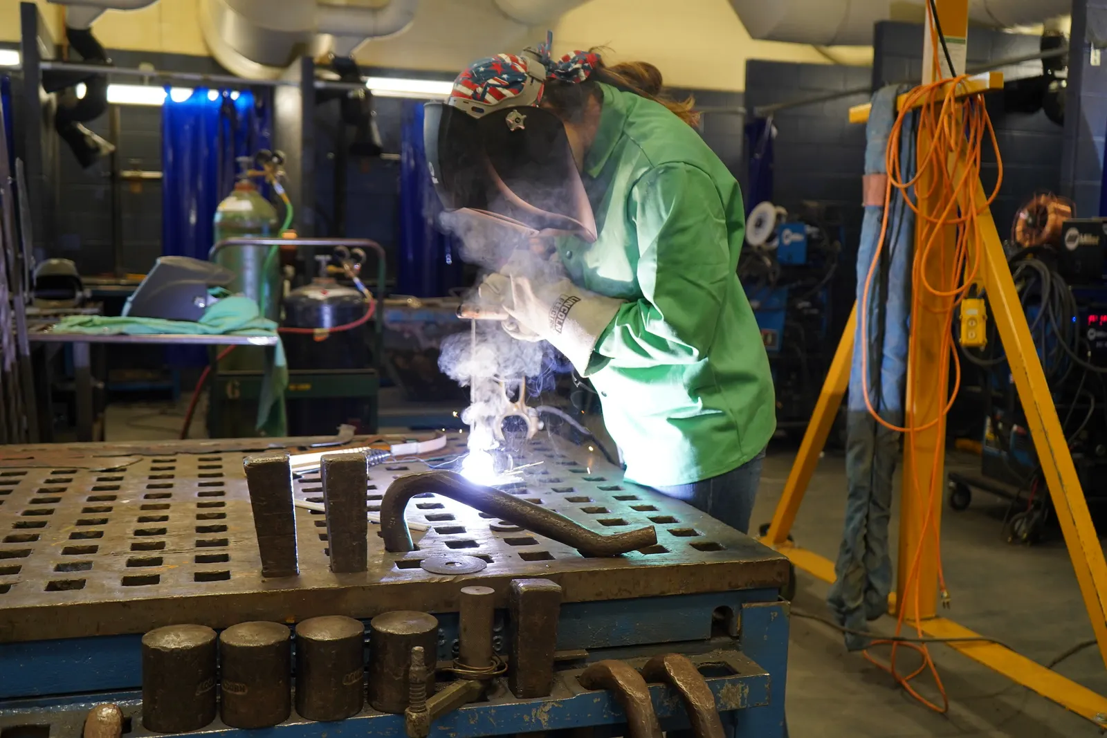 A student wearing a welding helmet and heavy gloves uses a welder on a metal piece in a workshop.